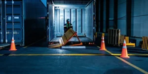 Truck driver assessing fallen pallets in a semi-trailer with orange safety cones and spilled freight