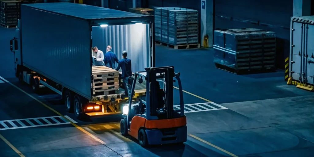 Tractor-trailer at freight yard with workers restacking shifted pallets using a forklift for emergency load correction